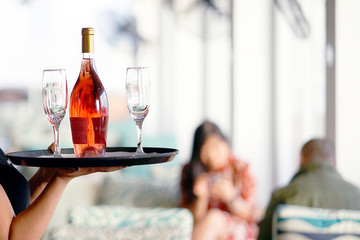 A young woman bartender carries a bottle of red wine with glasses on a tray to the client of the hotel restaurant. The concept of service.