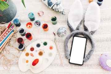 bunny ears and phone with empty screen ang egg and colorful paint on rustic background flat lay. space for text. creative easter image. greeting card mock-up. happy easter concept