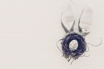 happy easter concept. bunny ears and stylish egg in nest on white wooden background flat lay with space for text. creative easter image. season's greeting card mock-up