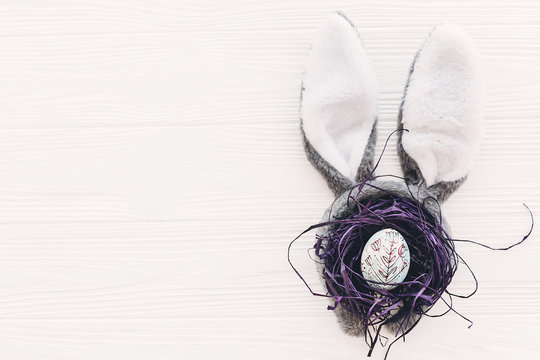 happy easter concept. bunny ears and stylish egg in nest on white wooden background flat lay with space for text. creative easter image. season's greeting card mock-up