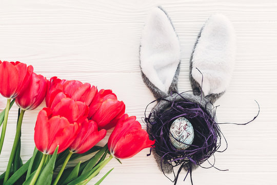 happy easter concept. bunny ears and stylish egg in nest  and tulips on white wooden background flat lay with space for text. creative easter image. spring season's greeting card mock-up