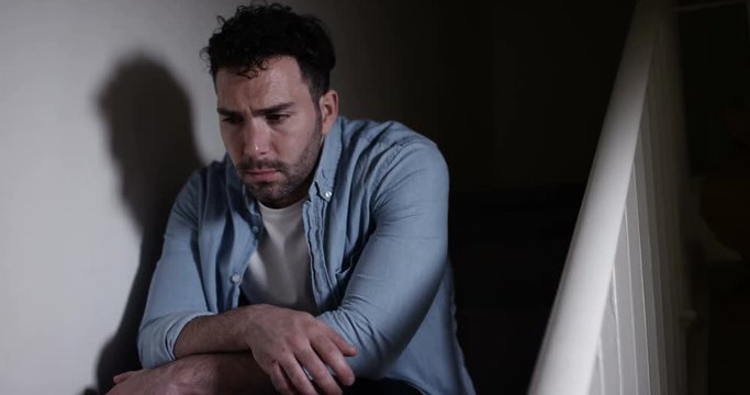 4K Depressed man sitting alone on the stairs at home. Loneliness concept, could be struggling with addiction or mental health issues or a victim of domestic abuse