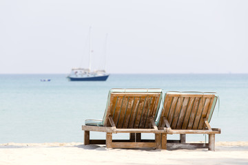 Couple of wooden beach chairs on a tropical sand beach overlooking the sea water and yacht. Thailand