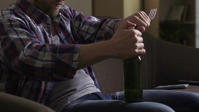 Single man sitting on couch and trying to open beer bottle with fork, despair