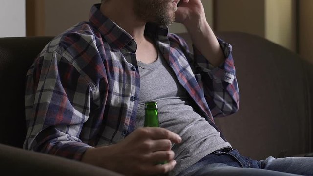 Angry addicted man sitting on couch, talking on cellphone and drinking beer
