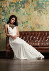 Beautiful brunette woman in luxurious evening gown sitting on sofa