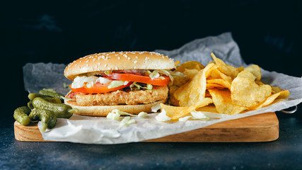 Chicken burgers on wooden tray. Two Homemade hamburgers, pickles and chips on a black metal background. Baking paper