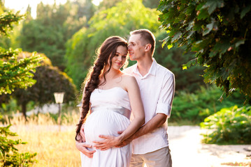Smiling young couple embracing in a green park on a sunny day enjoying pregnancy. Parents to be can't wait to welcome their baby. Devoted husband and wife showing of her pregnant belly.