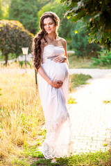 A pregnant woman in a white dress hugs her stomach and smiles in the park, the sun shines and the green grass. Concept of a happy woman and motherhood.