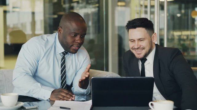 Two business colleagues looking at laptop computer and discussing their projects in modern office with glass walls. Bearded businessman and his partner sitting at table and talking