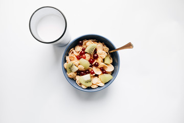 Delicious crispy cornflakes with kiwi pieces and cranberries in bowl, glass of milk on white background, closeup, healthy breakfast