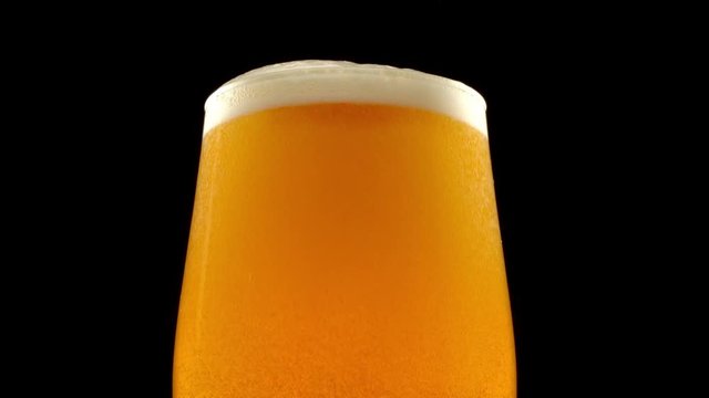 Glass of beer turns slowly around its axis. Beer bubbles rise to the beer foam. Black background. 