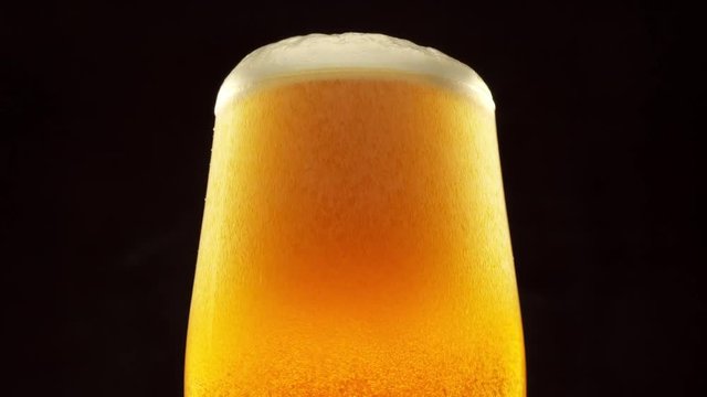 Glass of beer turns slowly around its axis. Beer bubbles rise to the beer foam. Black background. 