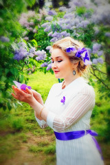 Beautiful blonde young girl in white dress with purple artificial butterflies in hands against blooming white and purple lilac