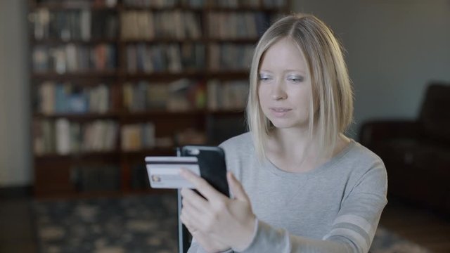 Girl buys online shopping at home with card and phone. Smiling young happy woman type entering master card number books sits on sofa indoors holding smartphone. Easy e-commerce transaction 4k