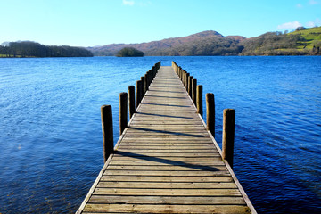 Obraz premium very long wooden symetrical beautiful wooden jetty, jutting out from the centre of the image into a calm blue lake with hills of forest and meadows in background