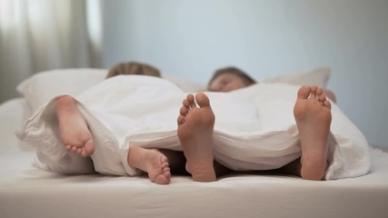 Girl touching feet of her boyfriend under duvet, romantic moments bed, closeness