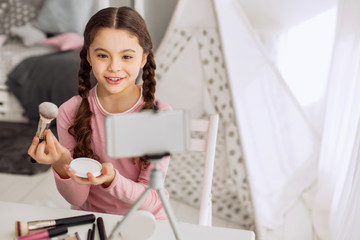Detailed review. Pleasant cheerful pre-teen girl making a review of makeup brushes and her new powder while filming it on her phone