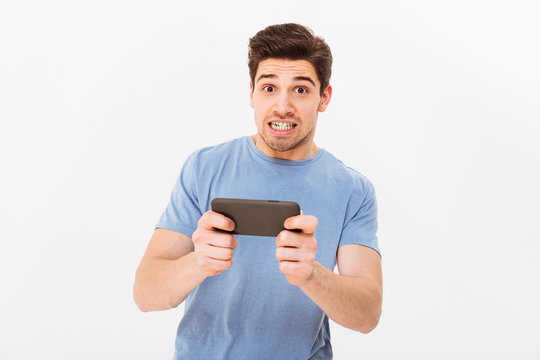 Adult game boy in casual t-shirt with tension on face having mobile entertainment while playing on smartphone, isolated over white background