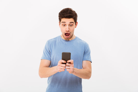 Photo of muscular guy 30s in casual t-shirt with excited facial expressions using mobile phone, isolated over white background