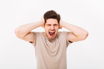 Disappointed man 30s grabbing his head or covering ears while screaming due to annoying noise, isolated over white background