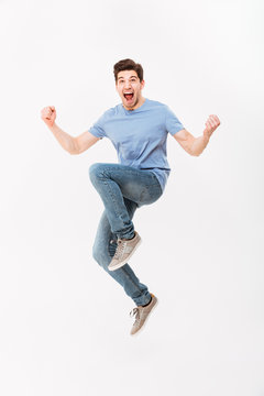 Full-length photo of excited man 30s in casual t-shirt and jeans levitating while expressing triumph with clenching fists, isolated over white background