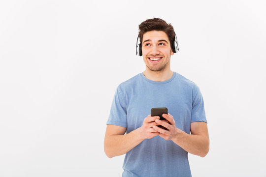 Caucasian smiling man in casual t-shirt listening to music via wireless earphones using smartphone, isolated over white background