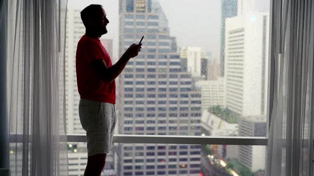 Young man using smartphone standing by window with city view, 4K
