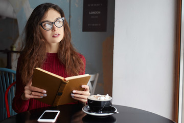 Young Beautiful Woman Reading Old Books, Using her Phone and Drinking Coffee or Cacao with Marshmallow. Relaxing atmosphere and Education.