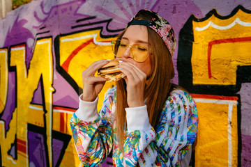 Charming hungry woman, enjoying eating a burger, closed her eyes. Dressed in stylish jacket and cap, in sunglasses. Standing against the wall with graffiti, outdoors.