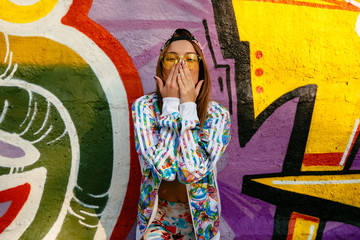 Attractive stylish girl covering her mouth by hands, looking at camera, wearing bright sportive suit and cap. Standing against the wall with graffiti.