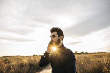 Content adult man with beard in stylish outfit speaking on phone against countryside in sunlight.