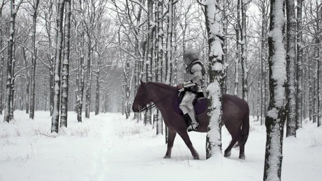 Portrait of medieval horseman wearing steel armor riding through winter forest, among snowy trees in slow motion. Reconstruction of ancient times
