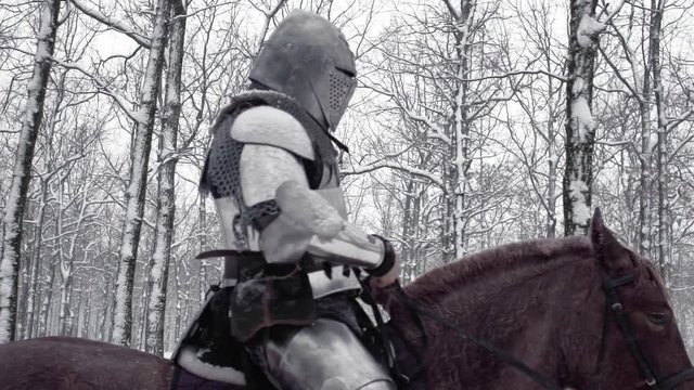 Strong lone gladiator armed with steel weapon riding on dark horse through winter forest, under snowfall in slow motion. Reconstruction of ancient times