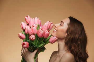 Image in profile of brunette female 20s with long hair holding and smelling bunch of pink flowers, isolated over beige background