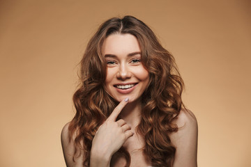 Elegant caucasian woman with long brown hair posing at camera with adorable smile, isolated over beige background