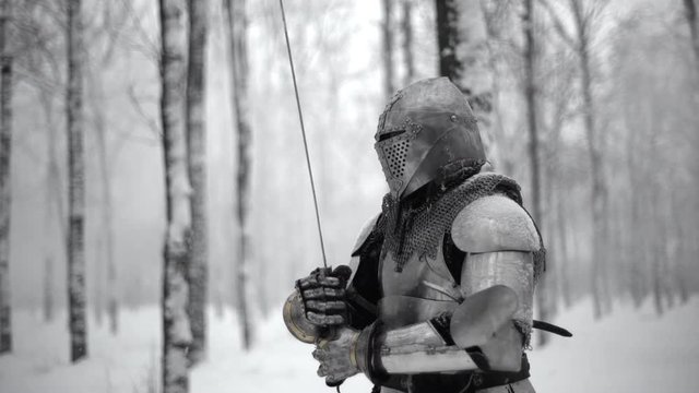 Portrait of brave warrior wearing steel armor and helmet preparing for swordfight, on battlefield in winter wood slow motion. Reconstruction of historical times