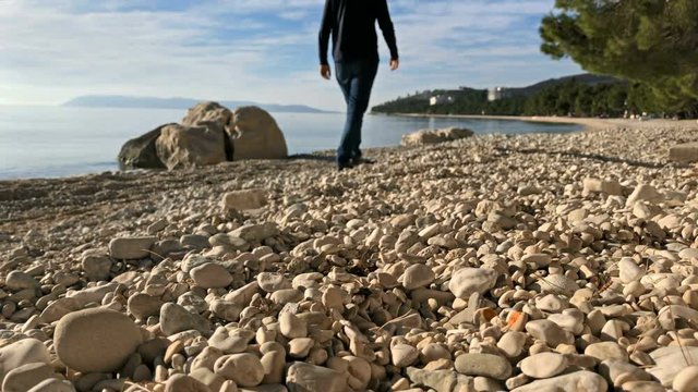 Young man in casual clothes walking on pebble beach near blue sea on sunny day slow motion