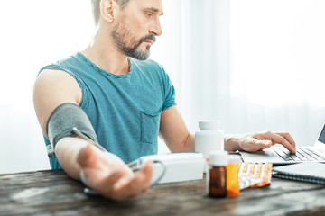 Bad news. Tired upset unshaken man sitting at home in the bright room by the table looking at the laptop and measuring pulse.