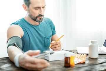 Different results. Embarrassing confused unshaken man sitting by the table measuring is pulse and looking at his notebook.