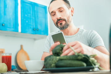 Best choice. Satisfied smart beardful man spending time in the kitchen taking vegetables and using the cellphone.