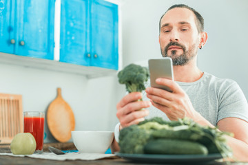 Check before eat this. Calm concentrated occupied man being in the kitchen looking at the cellphone holding a broccoli.
