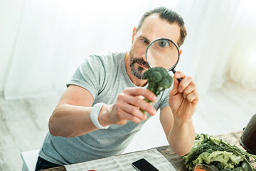 Amazing facts. Concentrated handsome beardful man spending time in the bright room holding a broccoli looking through magnifying glass.