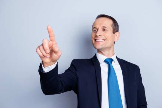 Cheerful, corporate, executive man in formal wear with tie having invisible screen, trying to touch a button with forefinger in front of him, standing over gray background