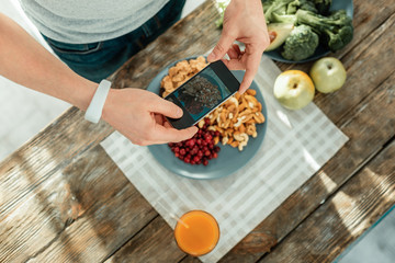 Good photo. Healthy creative concentrated man holding the smartphone in his hands making photo and standing near the table.