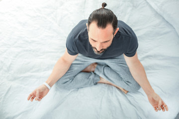 Way to karma. Tranquil calm handsome man sitting on the bed closing his eyes and having meditating.