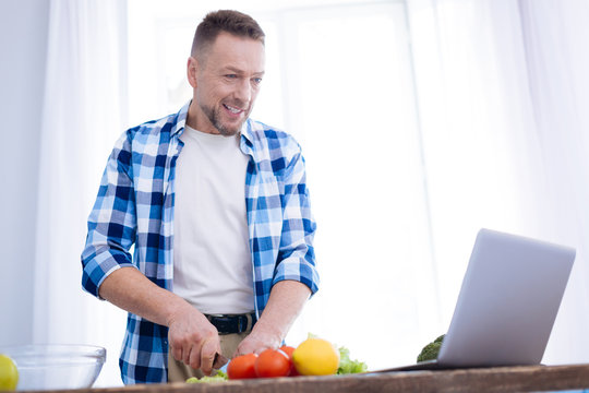 Healthy nutrition. Pensive musing gay man staring at screen while smirking and slicing vegetables