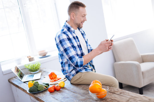 Food choice. Merry enthusiastic handsome man smiling while posing on surface and holding smartphone