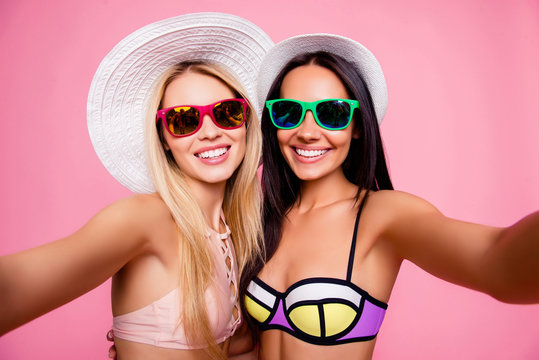 Self portrait of pretty, stylish, hot, cute, trendy tourists, ladies in swimsuits with beaming smiles shooting selfie together on front camera of smart phone, isolated on pink background
