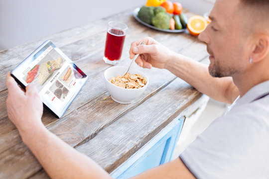 Homemade granola. Handsome stylish adorable man examining blog about biohacking while looking at screen and consuming granola